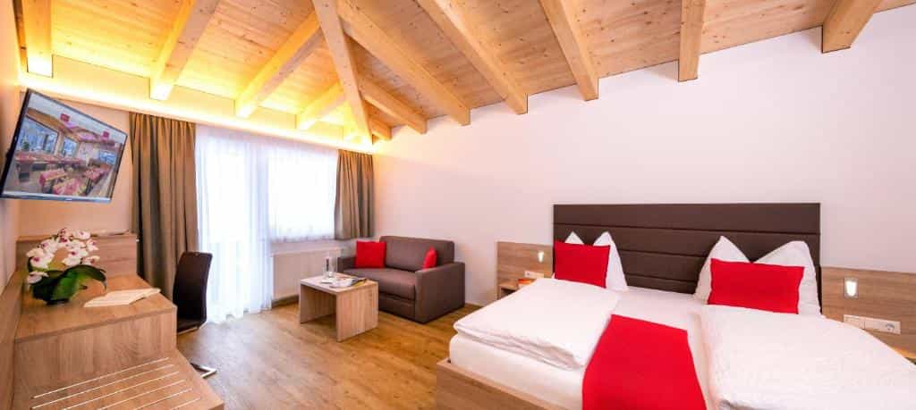 Hotel room with wooden beamed ceiling, modern bed with red accents, and a sofa.