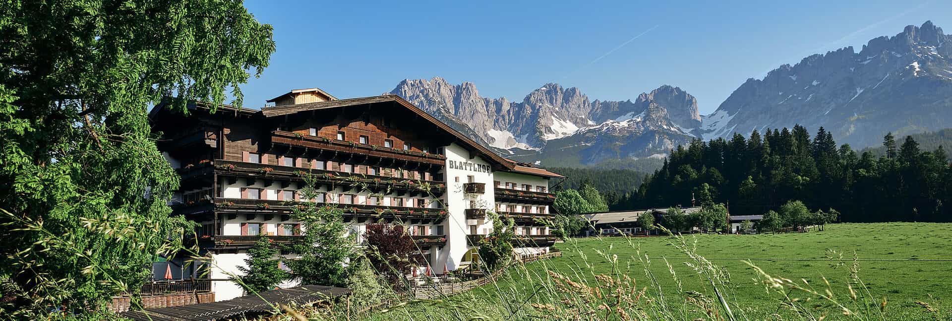 Hotel Blattlhof with wooden balconies set against jagged, snow-dusted alpine mountains.