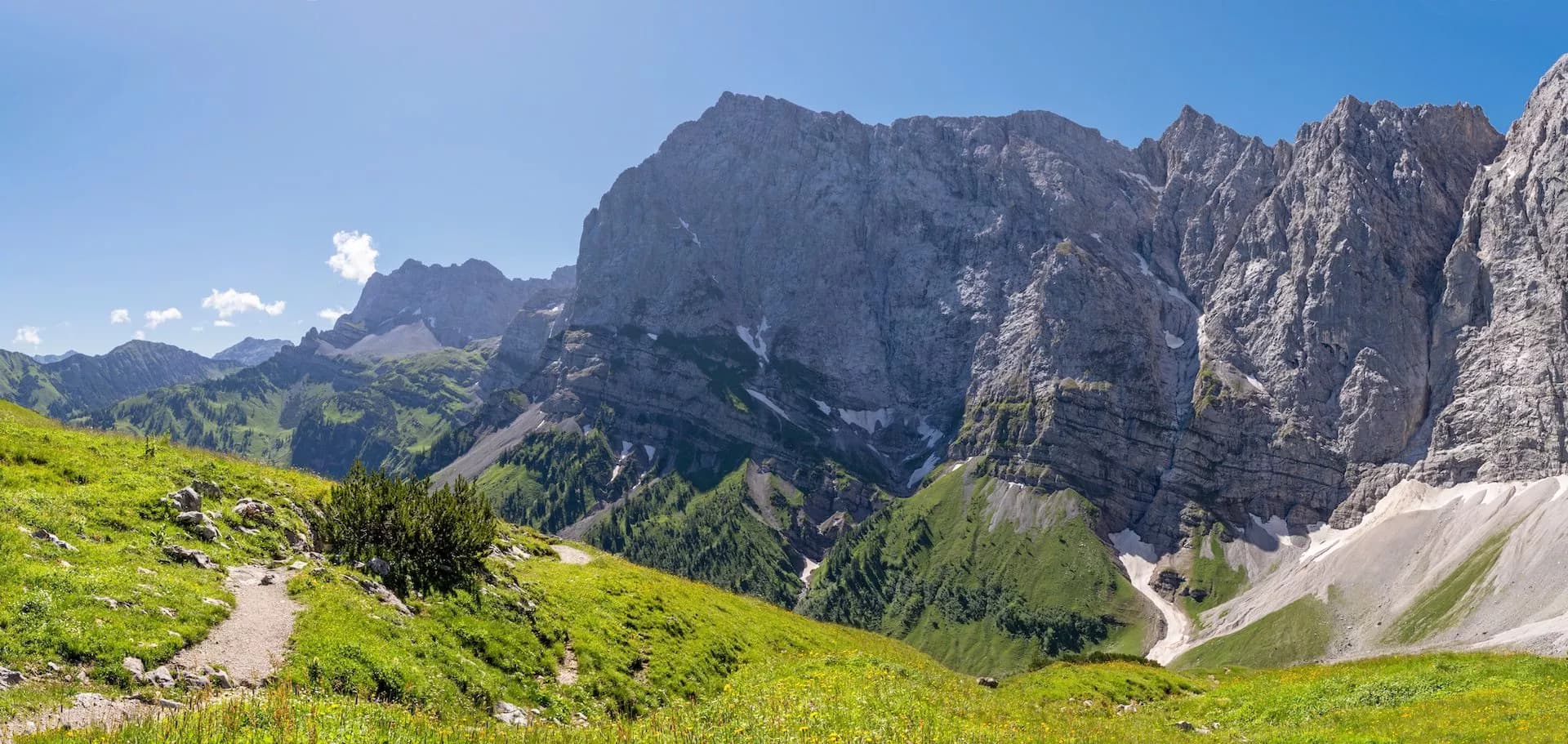 Hiking trail on grassy alpine meadow below massive gray mountains under blue sky