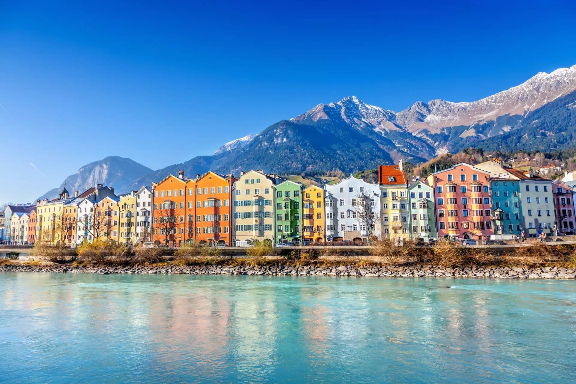 Colorful buildings along the river in Innsbruck with snow-capped mountains under a clear blue sky.