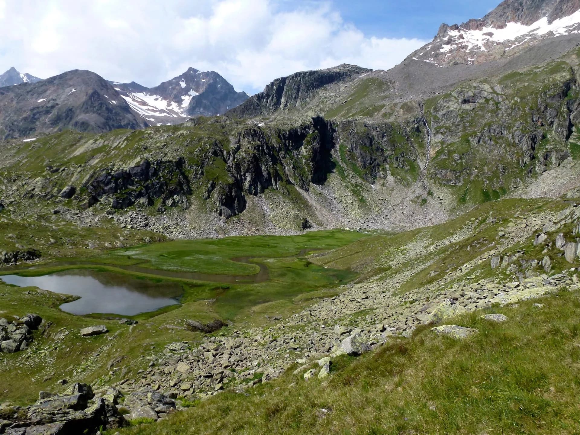 Alpine hiking trail with small lake, green meadow, and snow-capped mountains under a partly cloudy sky.