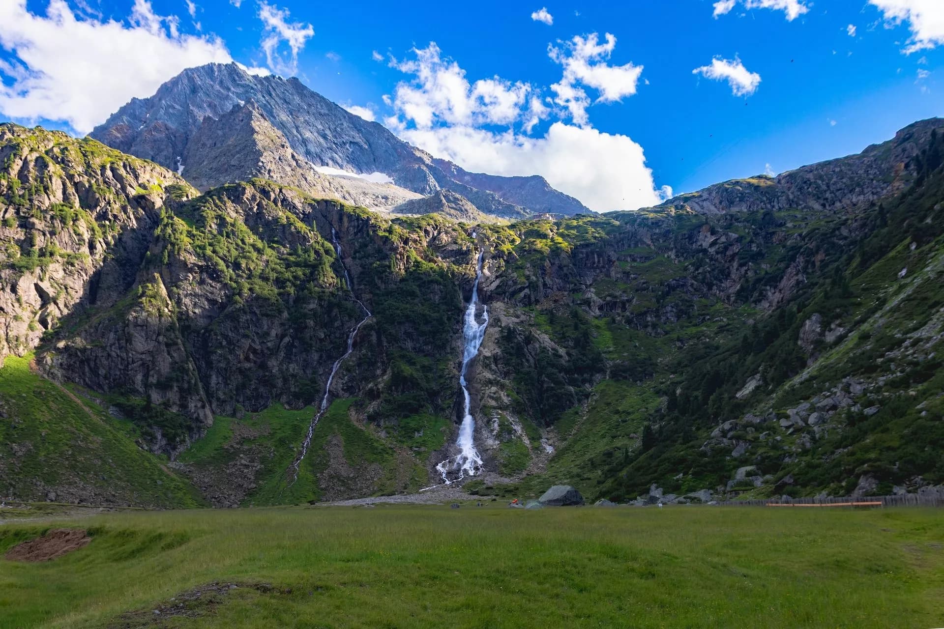 Waterfall cascading down steep, green mountainside under a bright blue sky with white clouds.