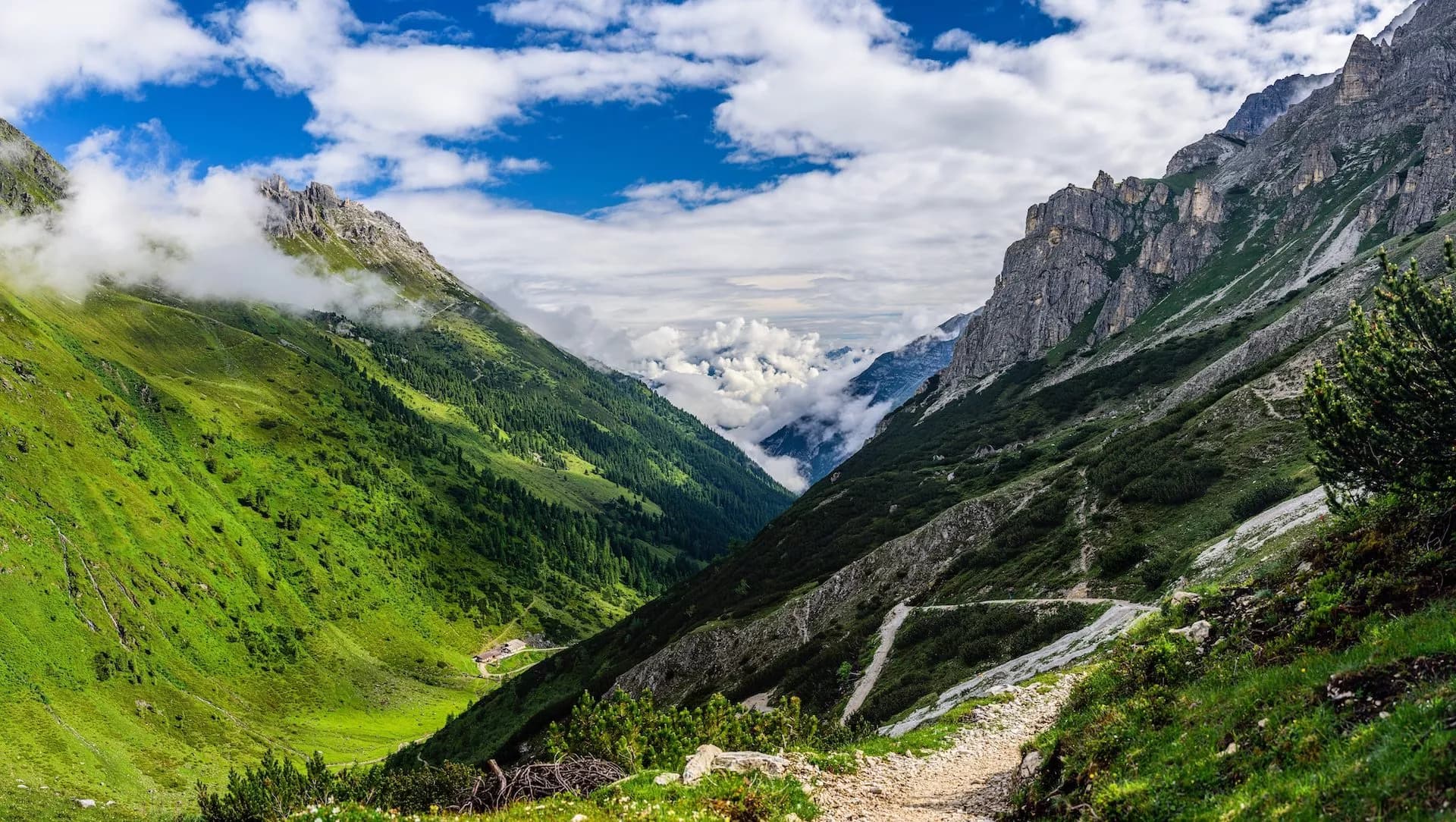 Hiking trail through a lush green alpine valley with rocky peaks and clouds below