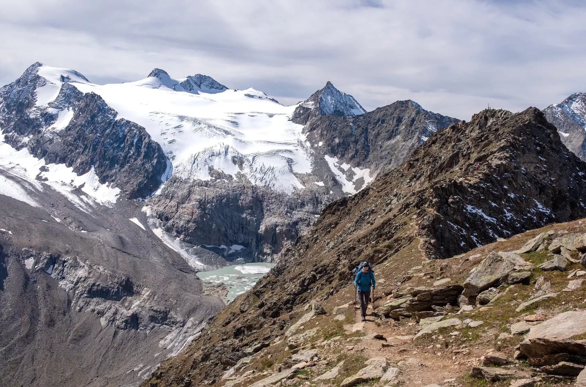 Hiker with poles on rocky trail below massive snow-capped glacier and glacial lake.