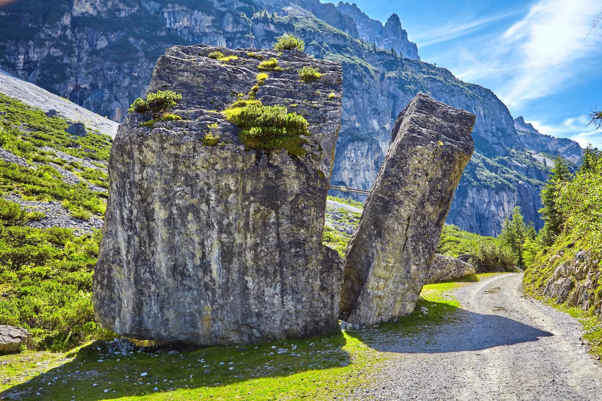 Split glacial erratic rock beside gravel path with steep green mountains under blue sky