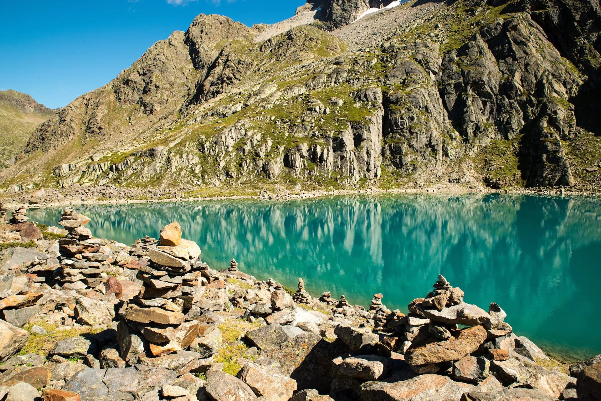 Rock cairns line the shore of a turquoise alpine lake beneath steep, rocky mountains.