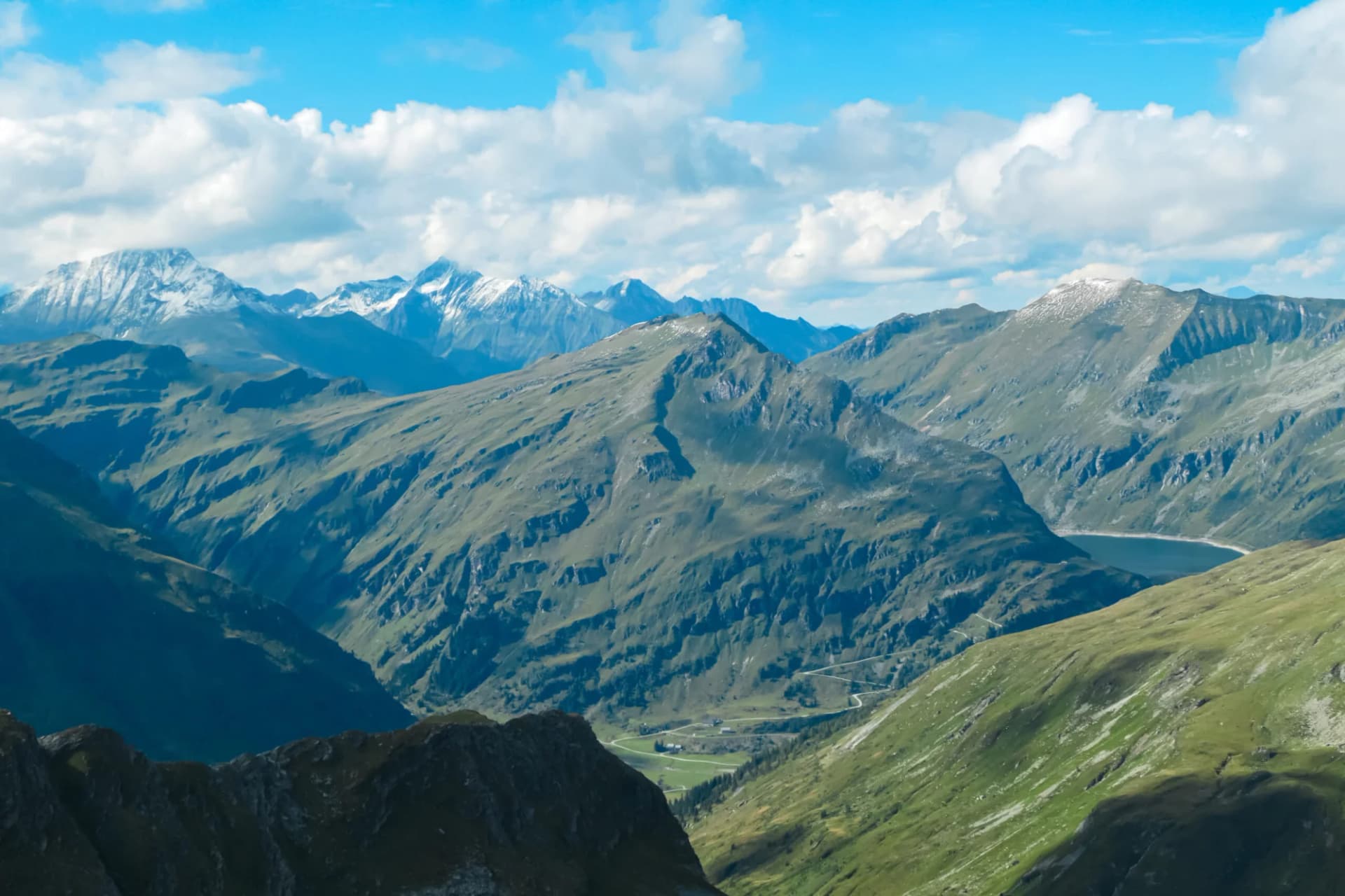 Alpine mountain range with green slopes, snow-capped peaks, and a reservoir under a blue, cloudy sky.