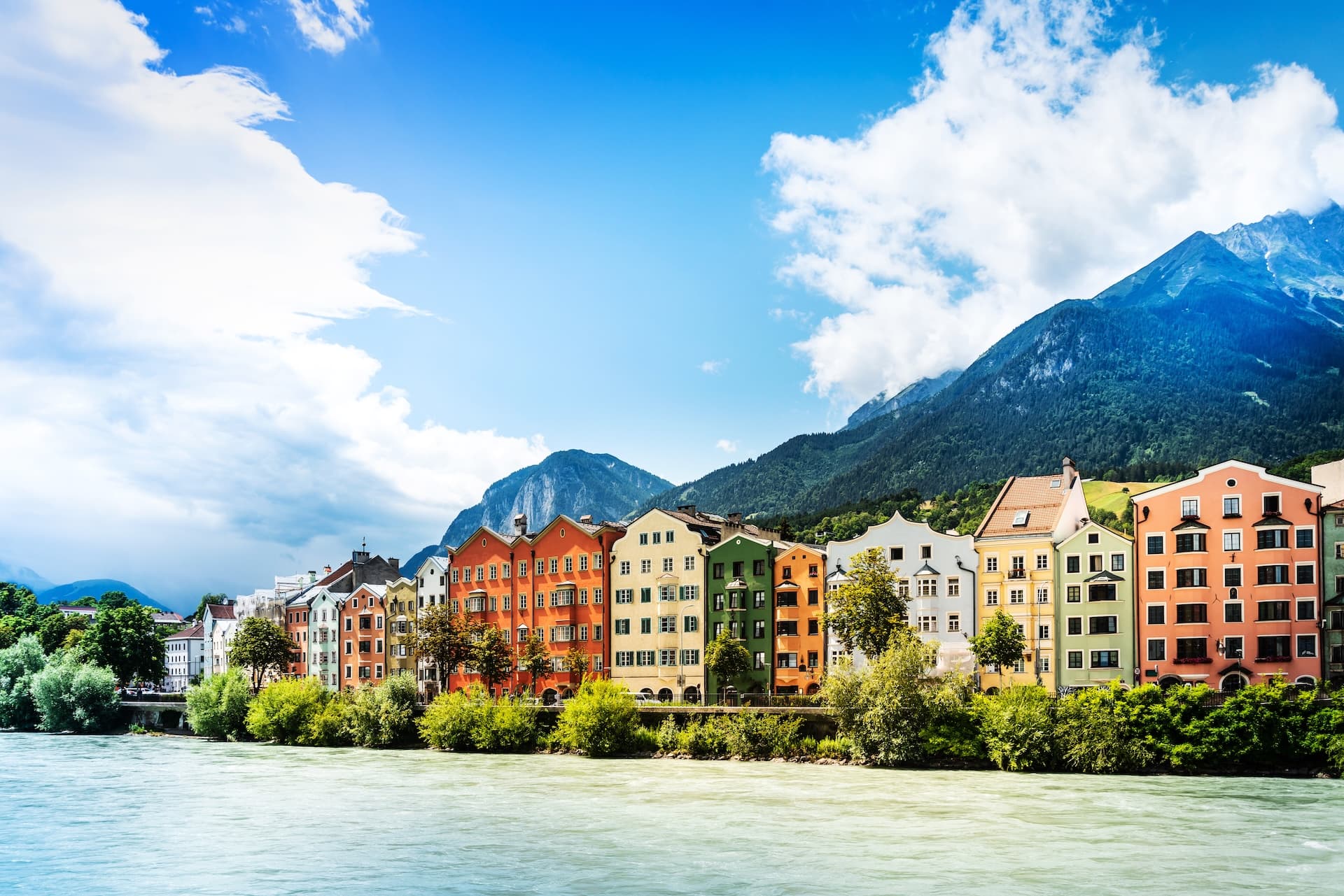 Colorful houses along the river with steep, forested mountains under a blue sky in Innsbruck.