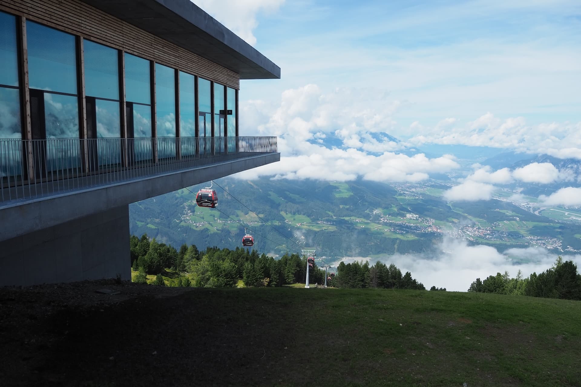 Gondola lift ascending past modern building overlooking green valley and mountains below clouds