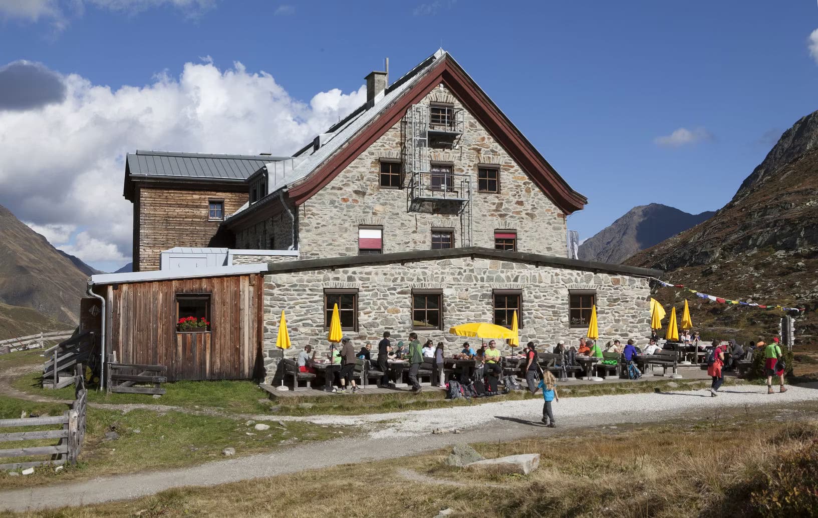 Franz-Senn-Hütte stone mountain lodge with hikers dining at outdoor tables under yellow umbrellas in the Alps.