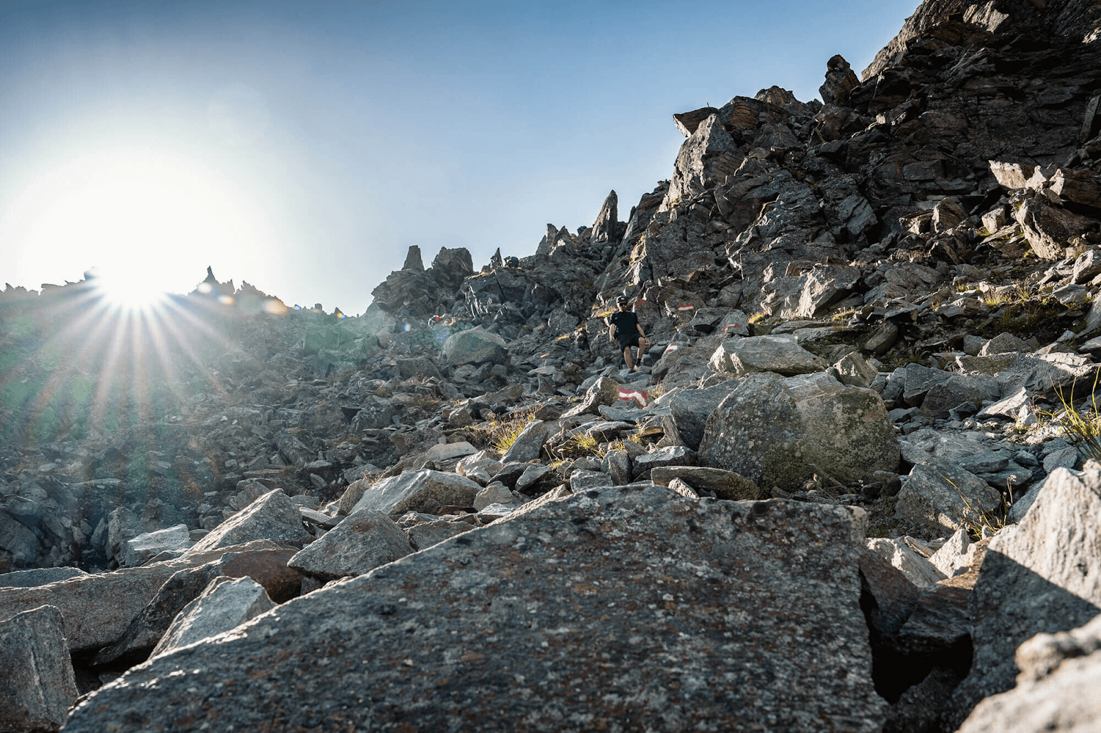 Hiker descending a rocky mountain trail in the Stubai Alps with bright sun flare.