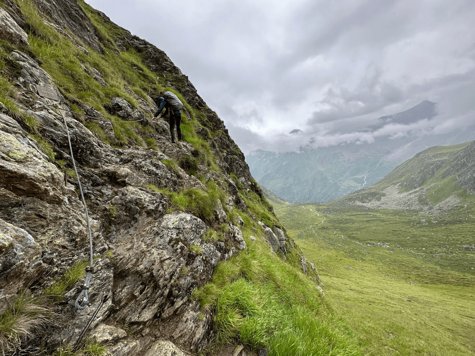 Hiker on a steep via ferrata trail in the Stubai Alps with a green mountain valley below.