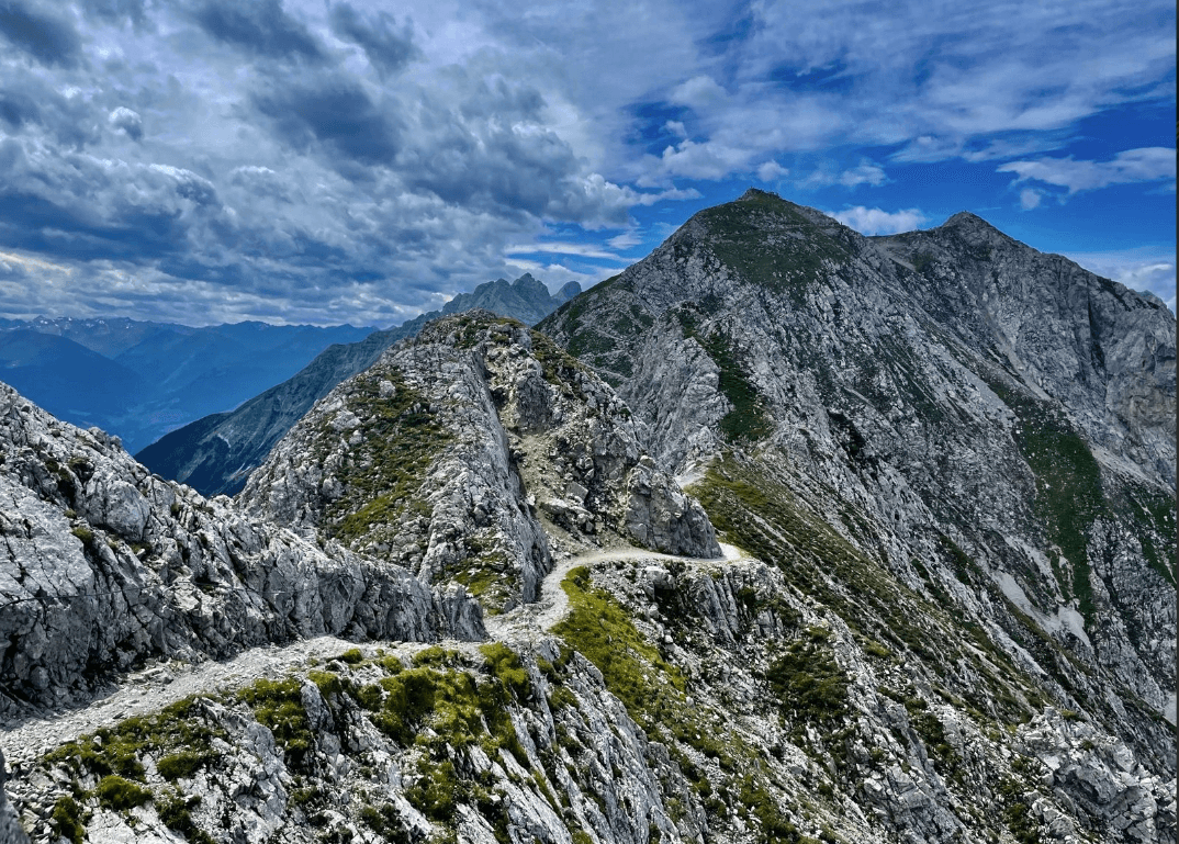 Hiking trail along a rocky mountain ridge with dramatic peaks under a cloudy sky.