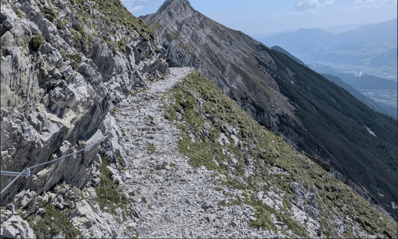 Rocky hiking trail with a safety cable along a steep alpine mountain slope.