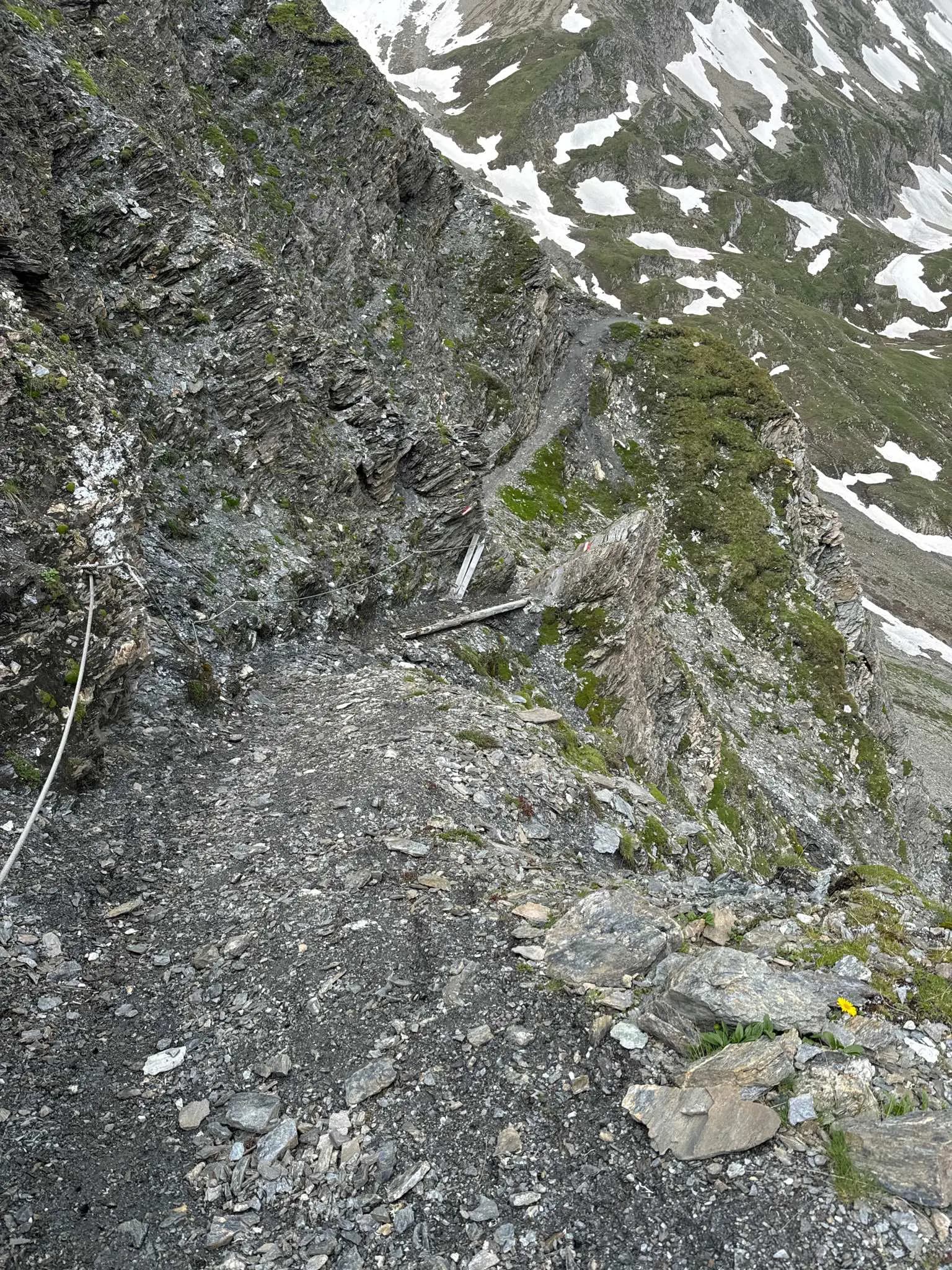 Narrow hiking trail on a steep rocky mountain slope with patches of snow and a safety cable.