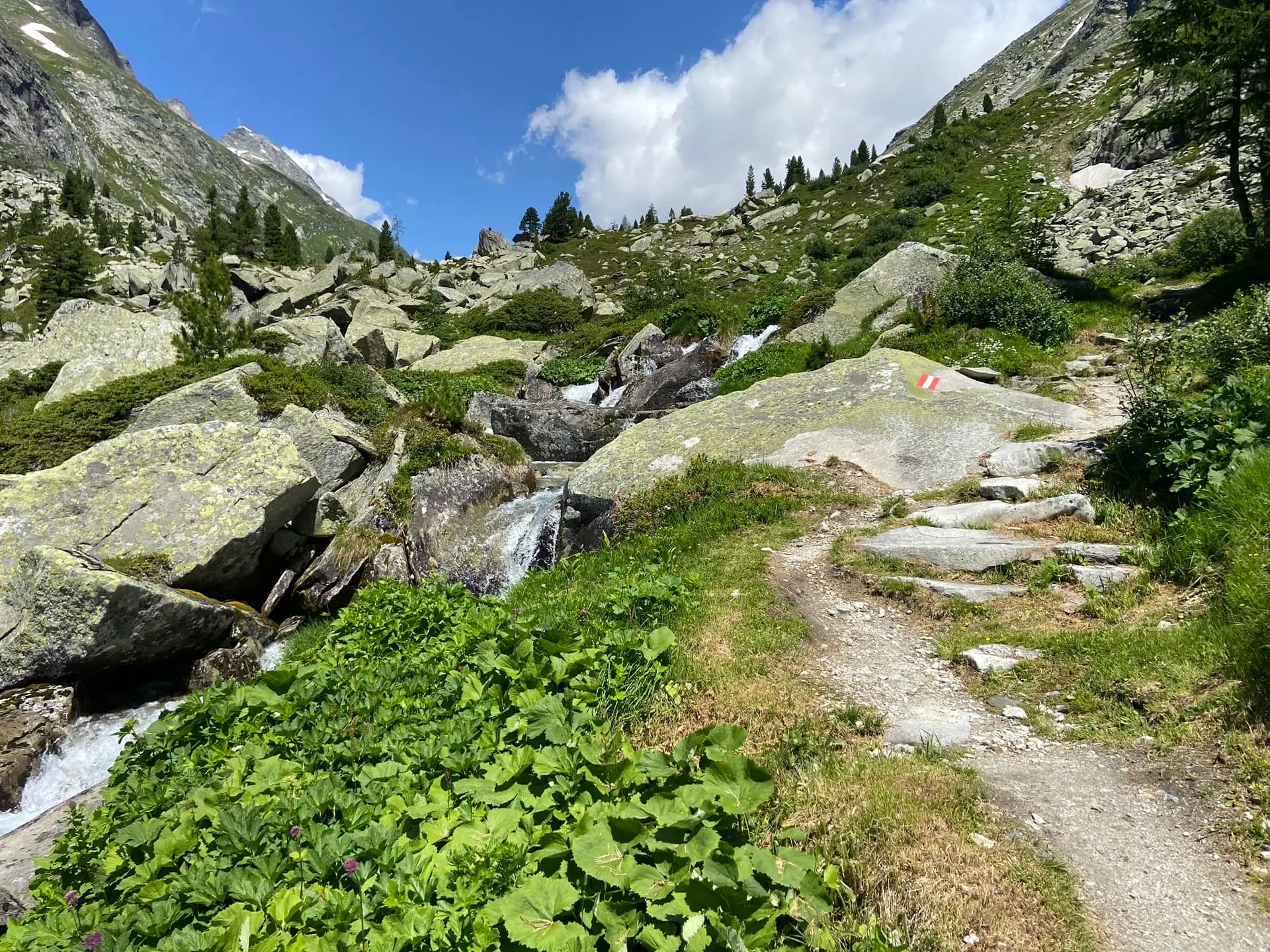 Hiking trail with red and white marker along a mountain stream in a rocky alpine landscape.
