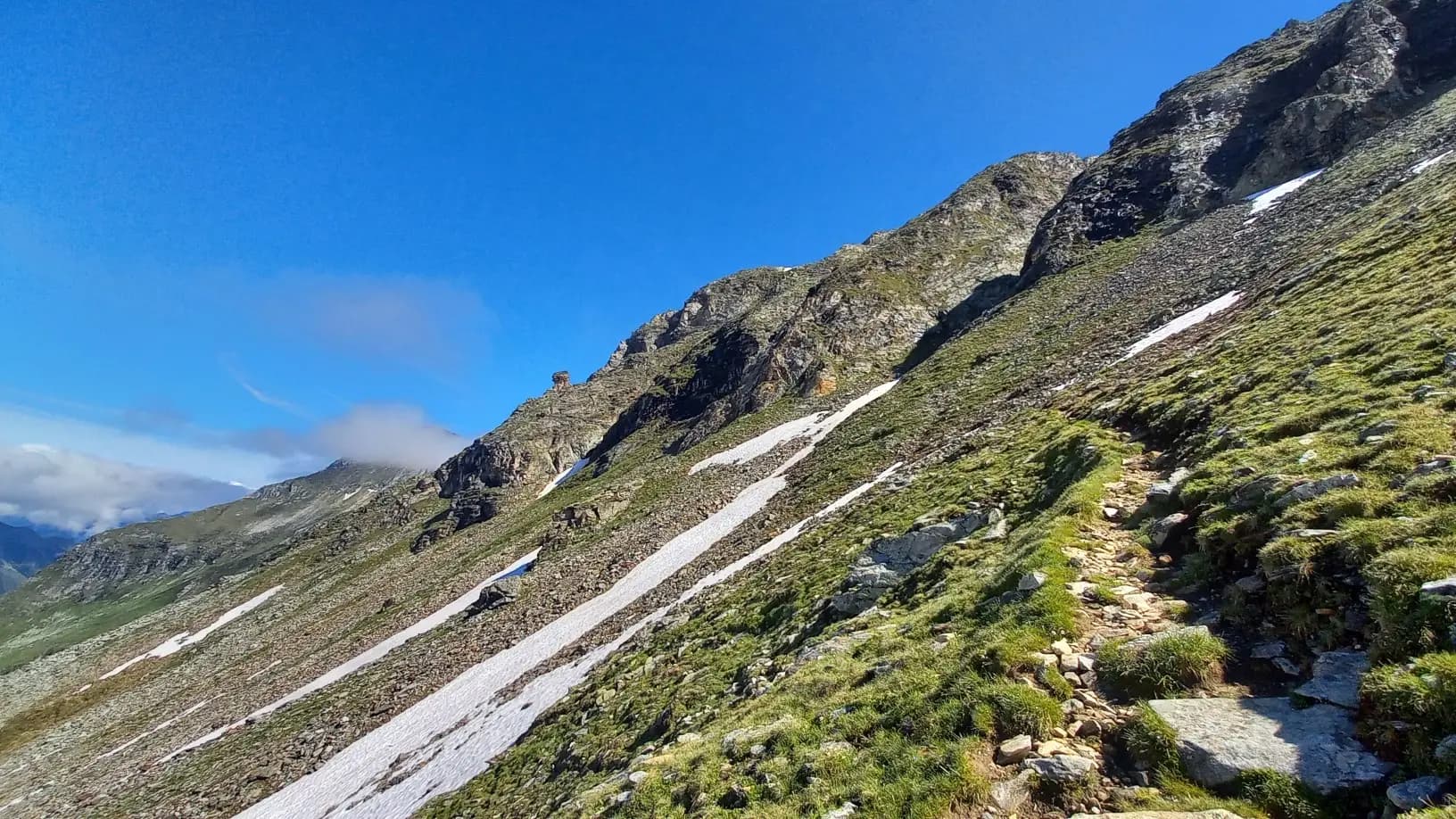 Hiking trail on a grassy mountain slope with patches of snow in the Hohe Tauern range.