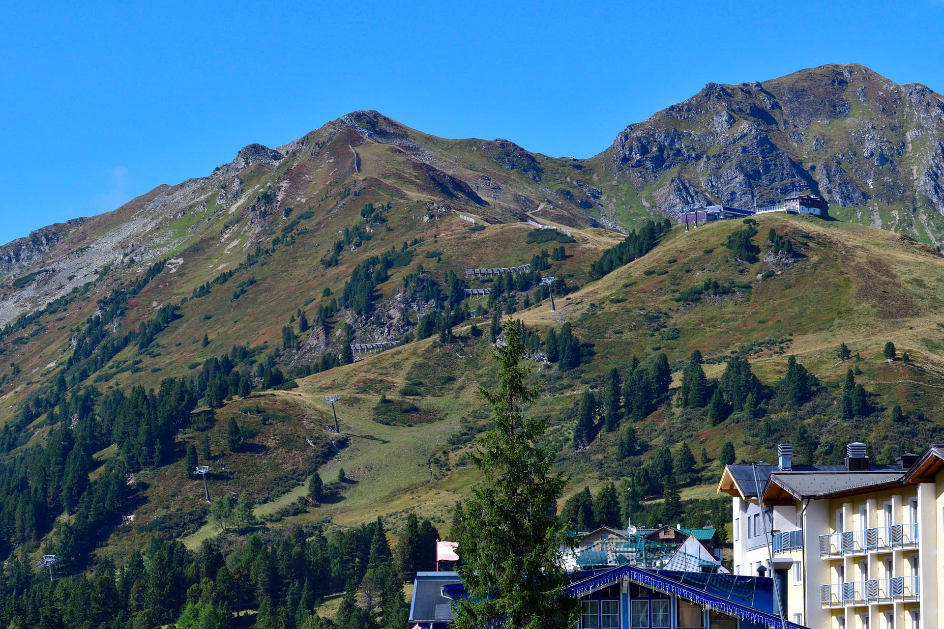 Alpine town buildings below grassy mountains with visible ski lift infrastructure under clear blue sky.