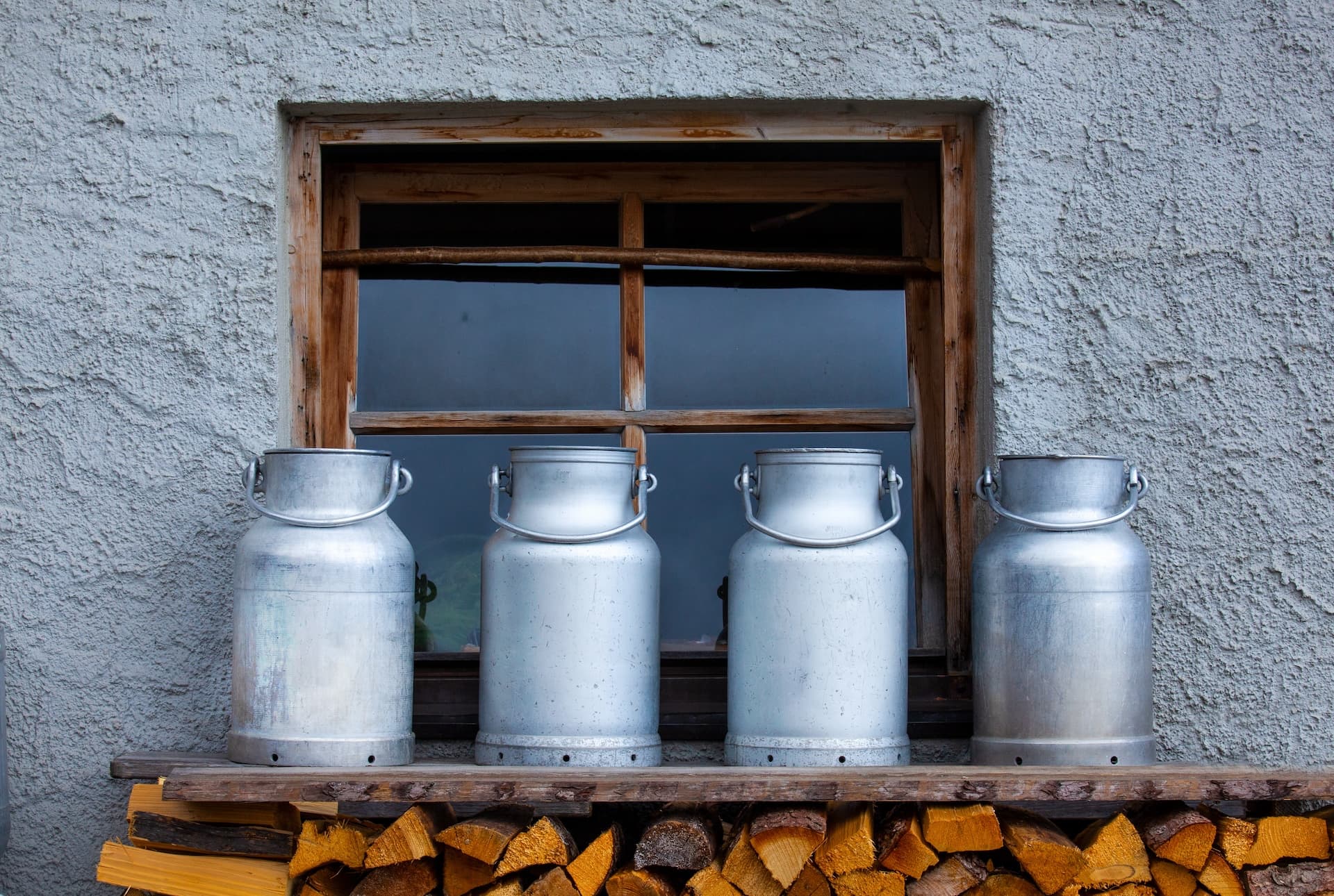 Four metal milk containers on a wooden shelf above stacked firewood against a stucco wall.