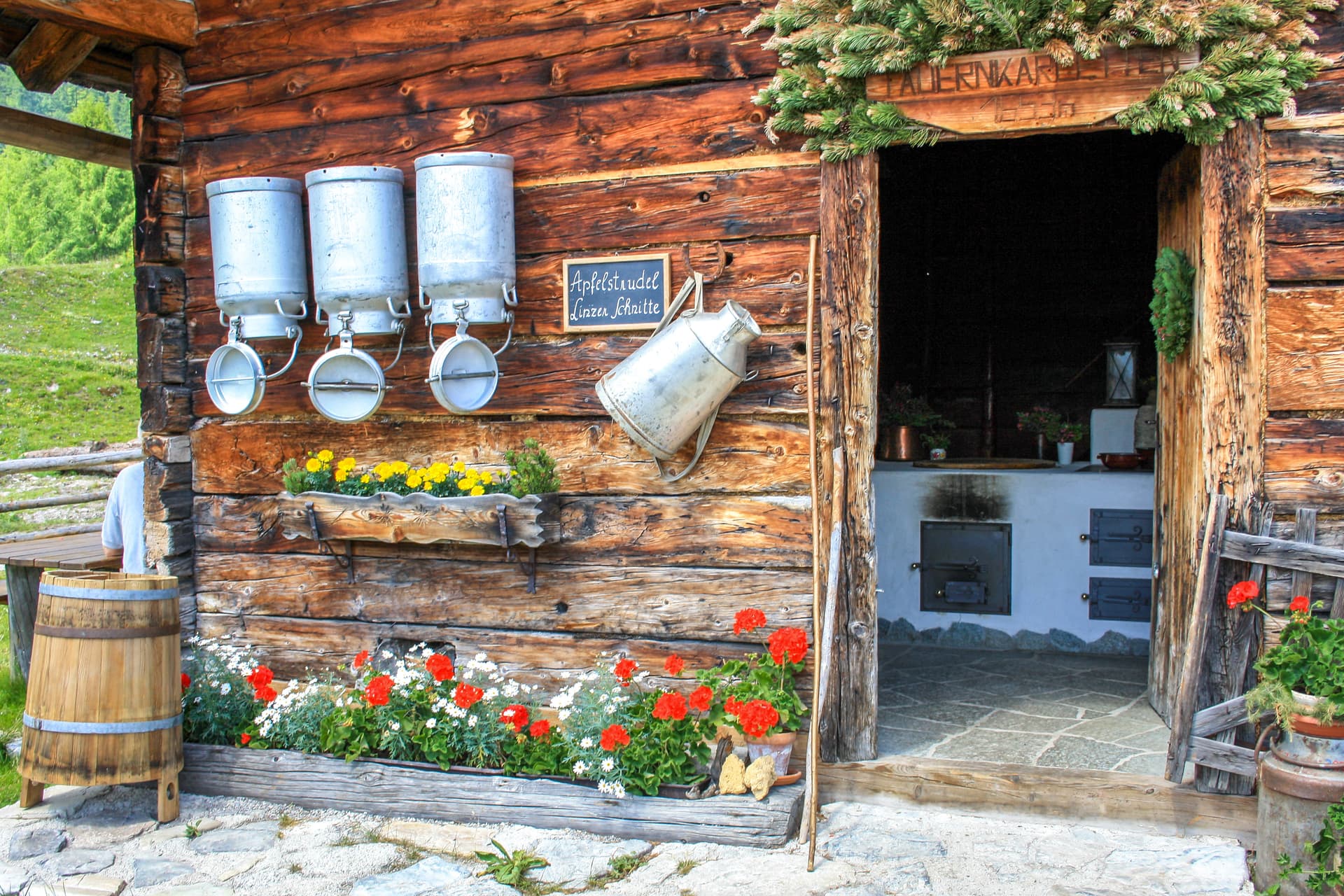 Rustic wooden cottage exterior in Austria with milk cans, flowers, and green mountain backdrop.