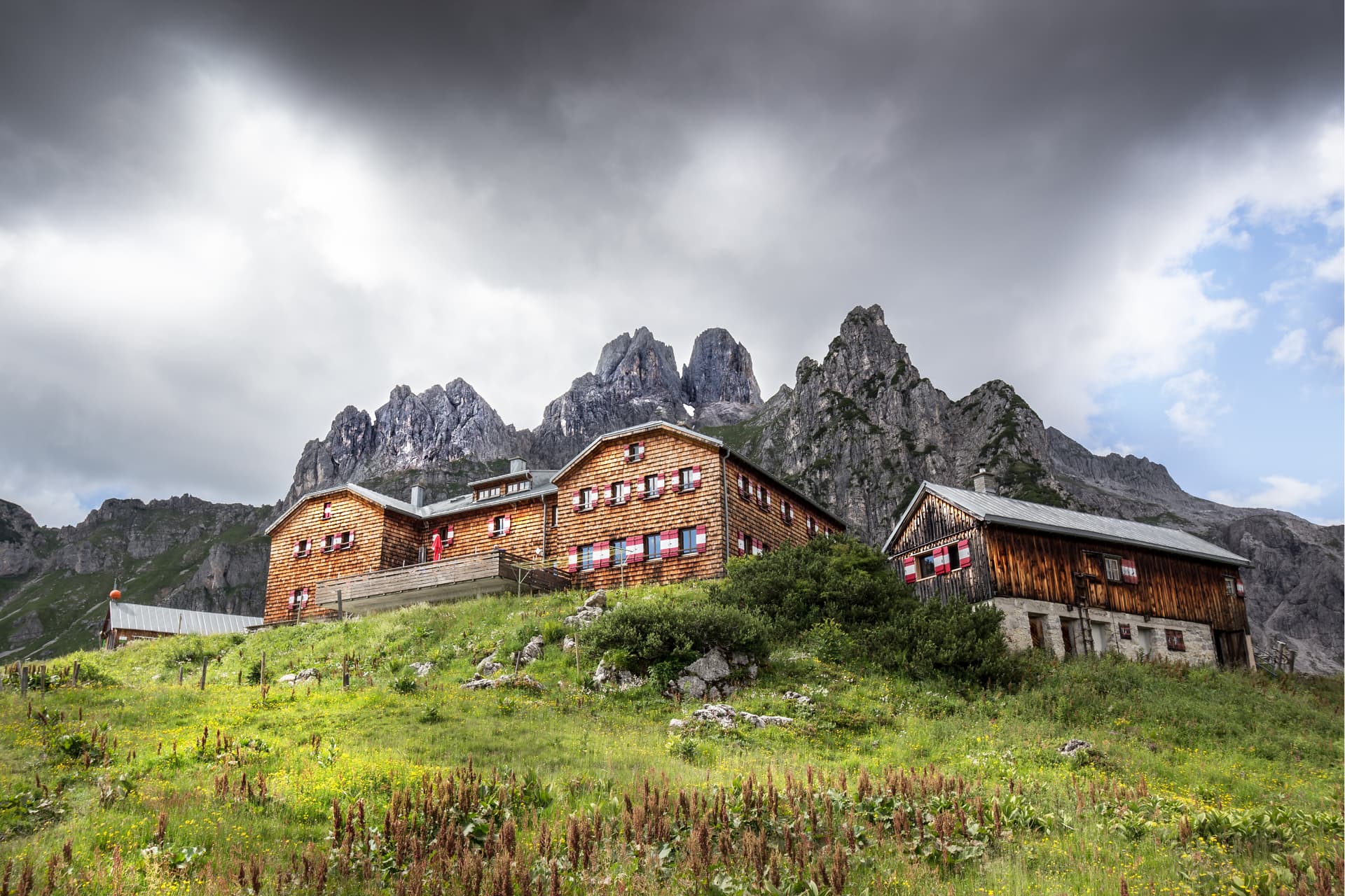 Wooden alpine hut nestled on a grassy slope below rugged, rocky mountains under a dramatic sky.