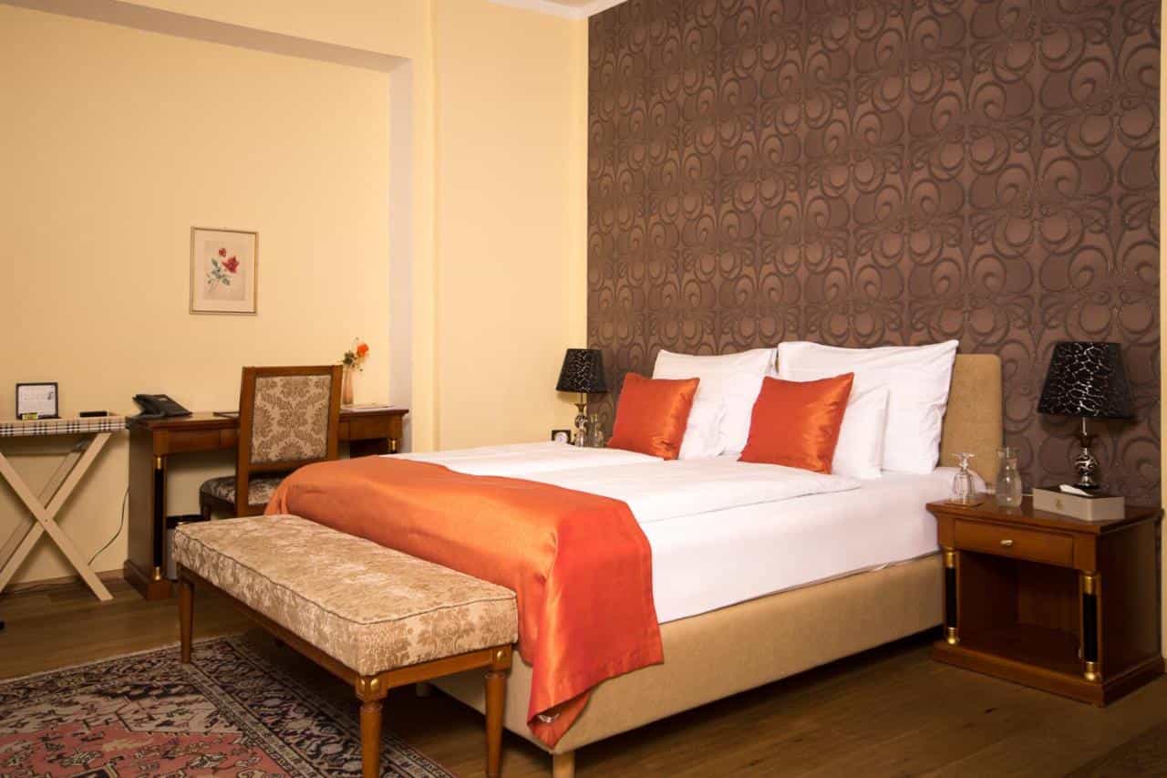 Bedroom with beige walls, white bedding, orange accents, and brown patterned wallpaper at Seehotel Grüner Baum.