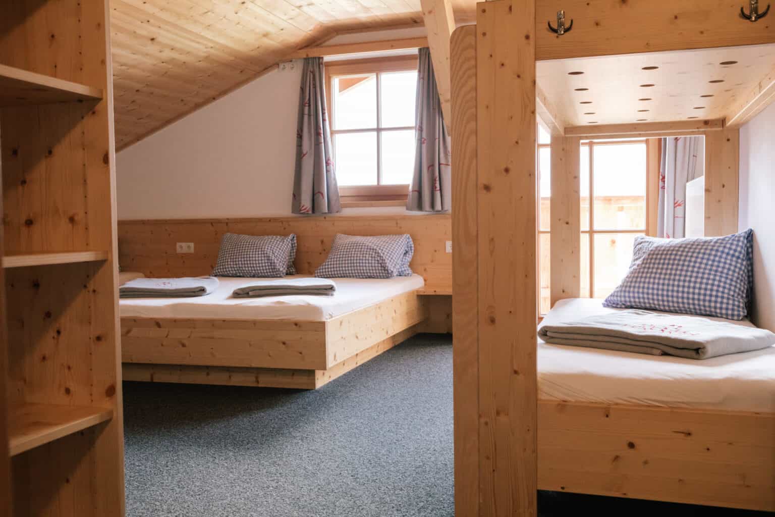 Wooden alpine room interior with beds, slanted wood ceiling, and bright window views.