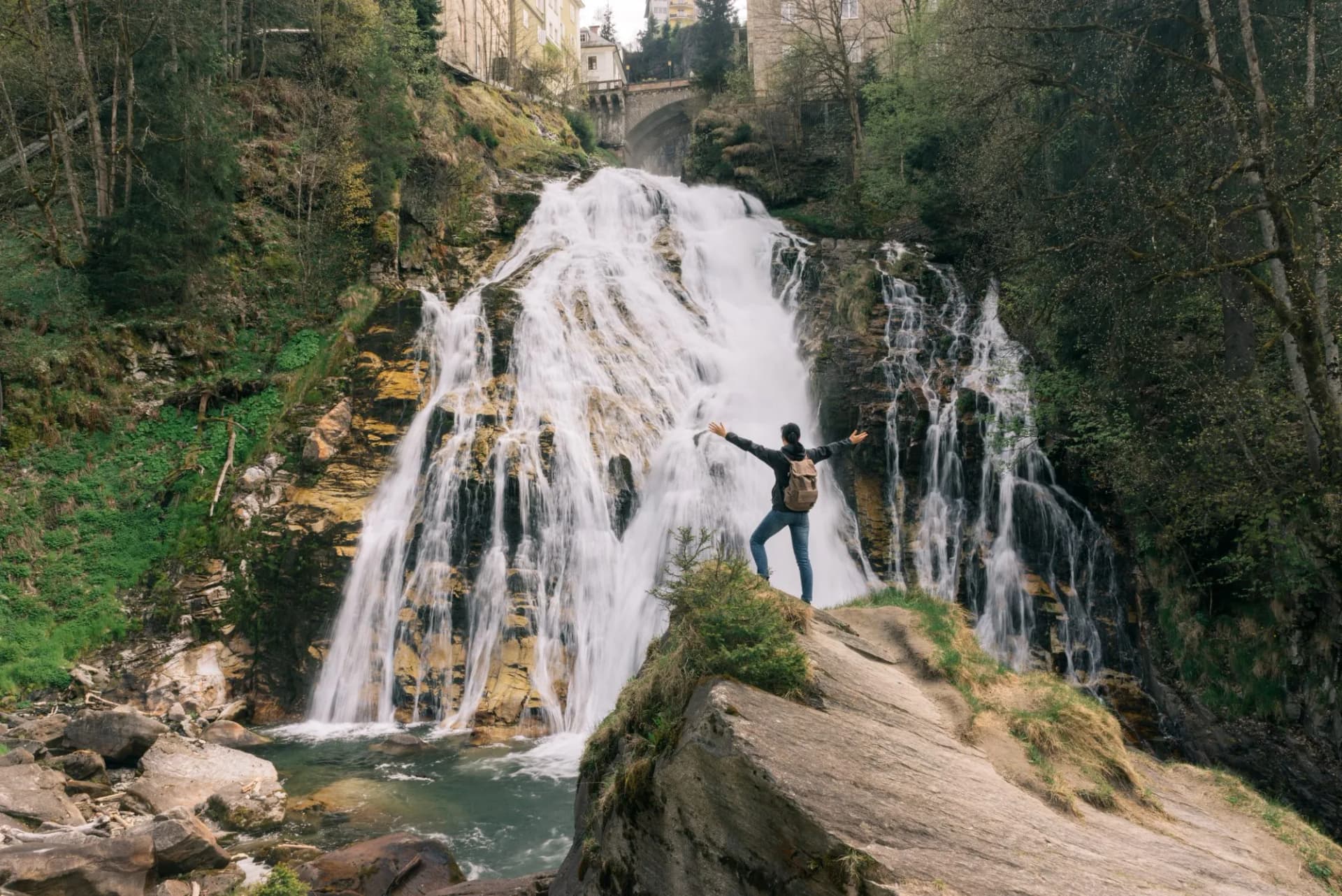 Hiker with arms spread before the Gastein Waterfall with buildings visible above the gorge.